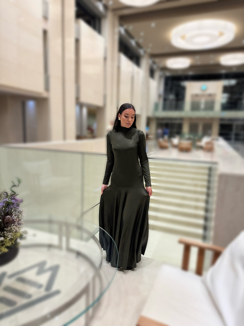 A woman in a dark olive green dress standing in a modern indoor setting with glass walls and stairs.