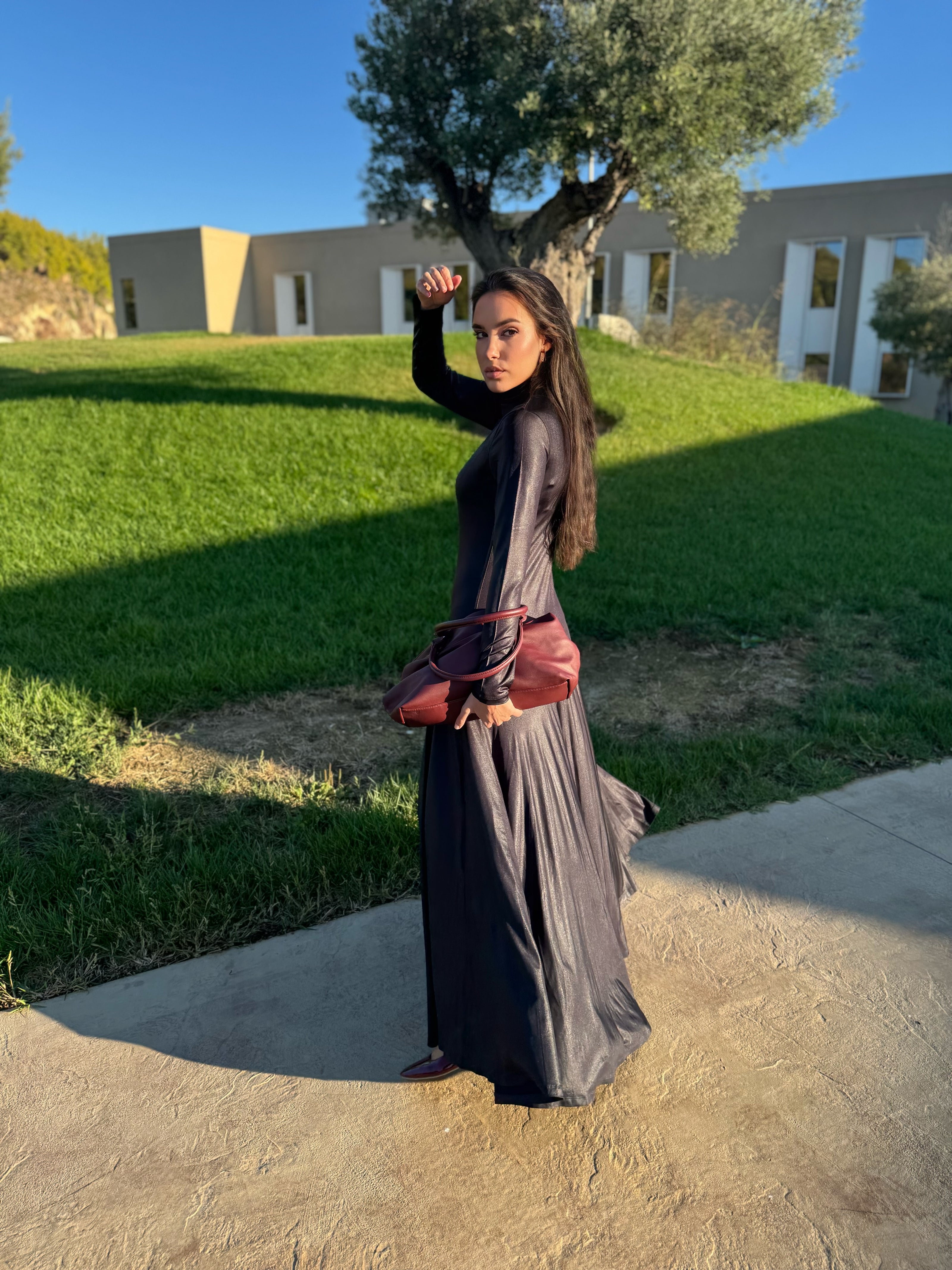 Woman in a long dark dress, which moves with her, holding a red purse outdoors with green grass and a building in the background.