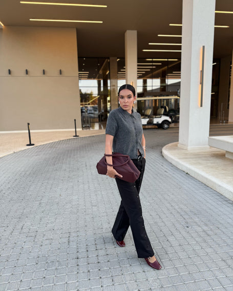 Stylishly dressed woman standing on a paved area with a building in the background wearing a bag