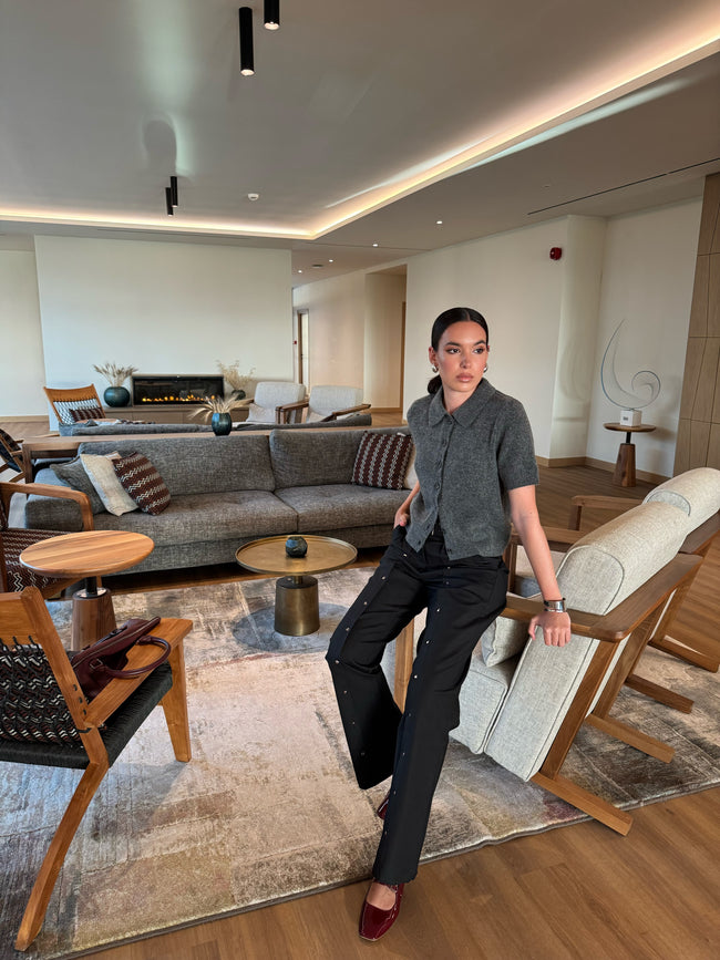 Woman standing in a modern living room with gray sofa and wooden chairs. She is wearing flurry and wide pants with silver drinkers along the full length of the pants.
